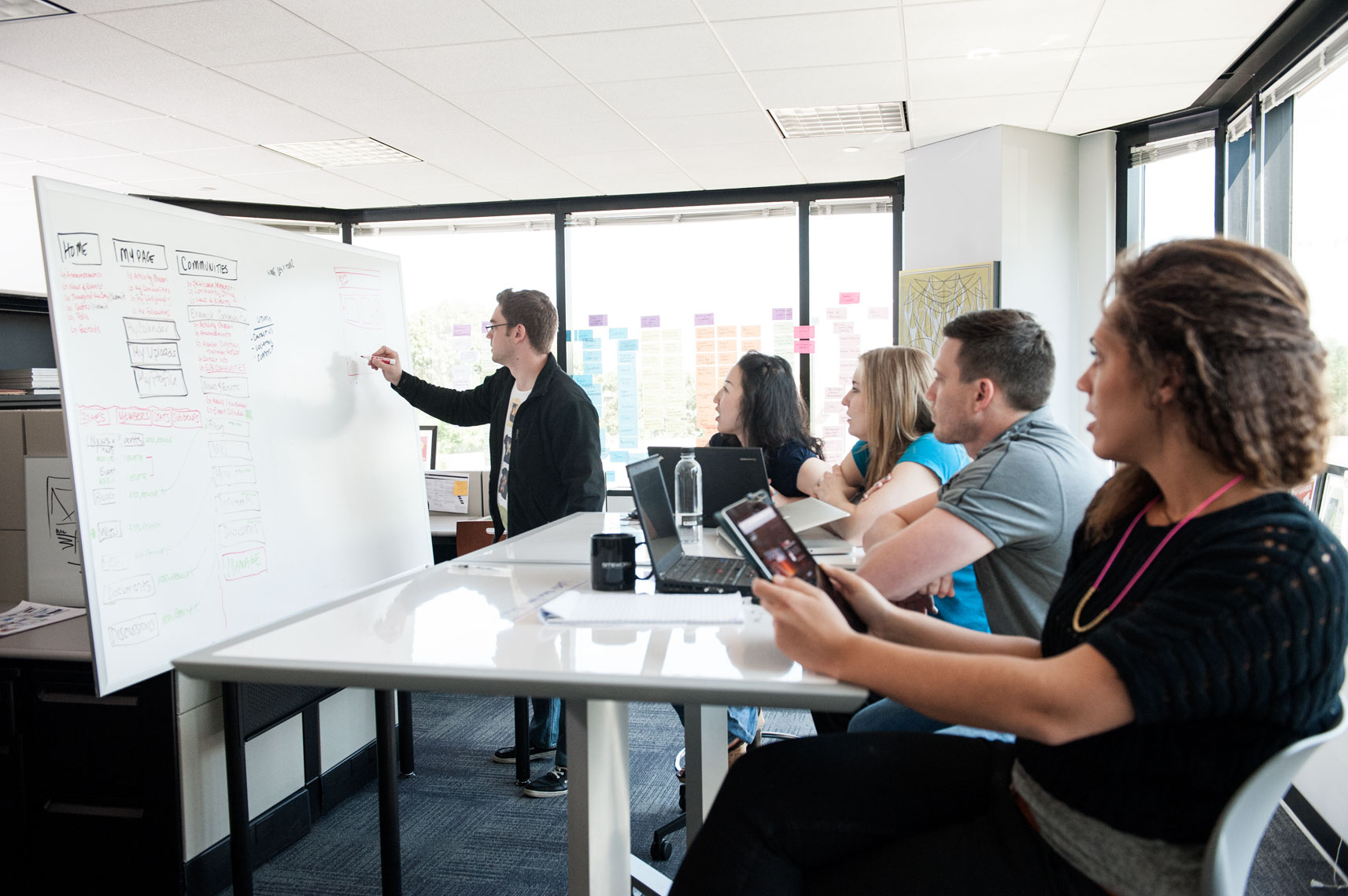 A man works at a whiteboard in front of a table of people with electronic devices
