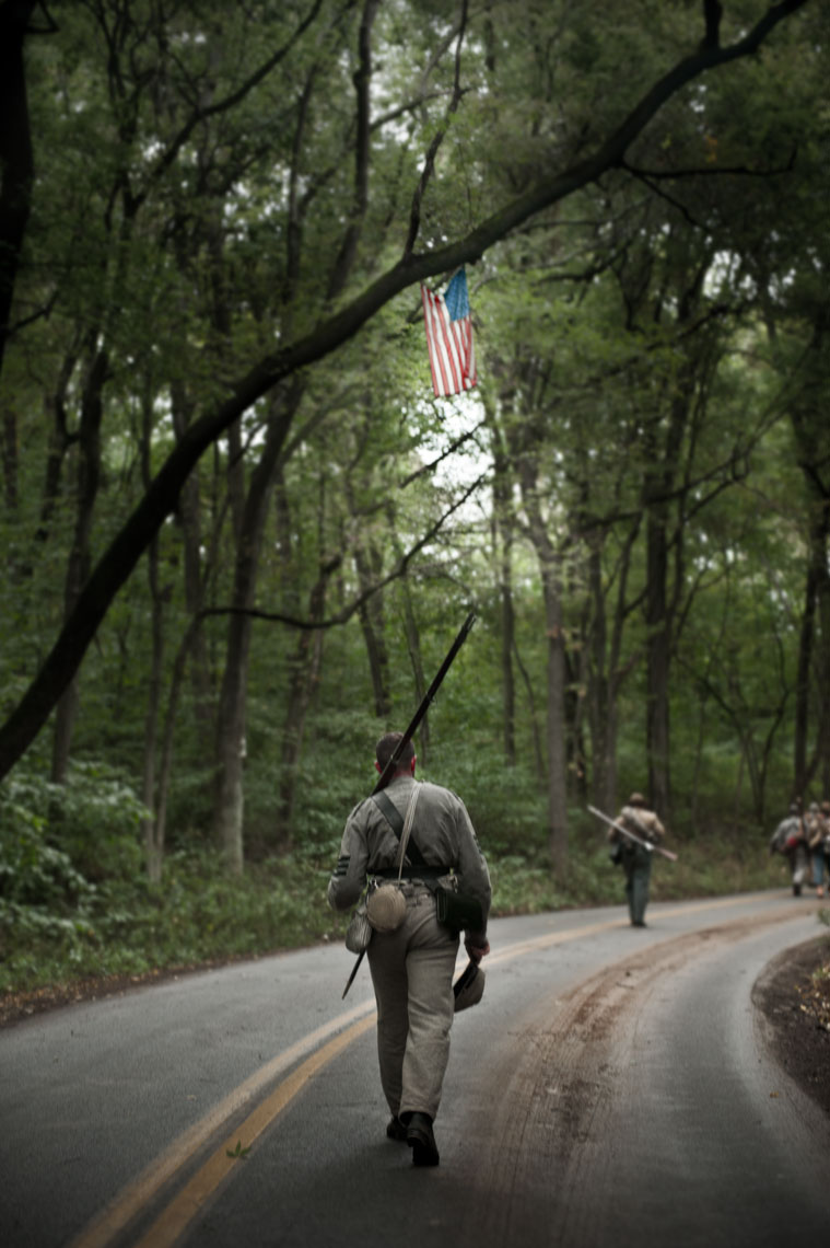 Civil War reenactors at Antietam: reenactors walk along an empty road beneath an American flag