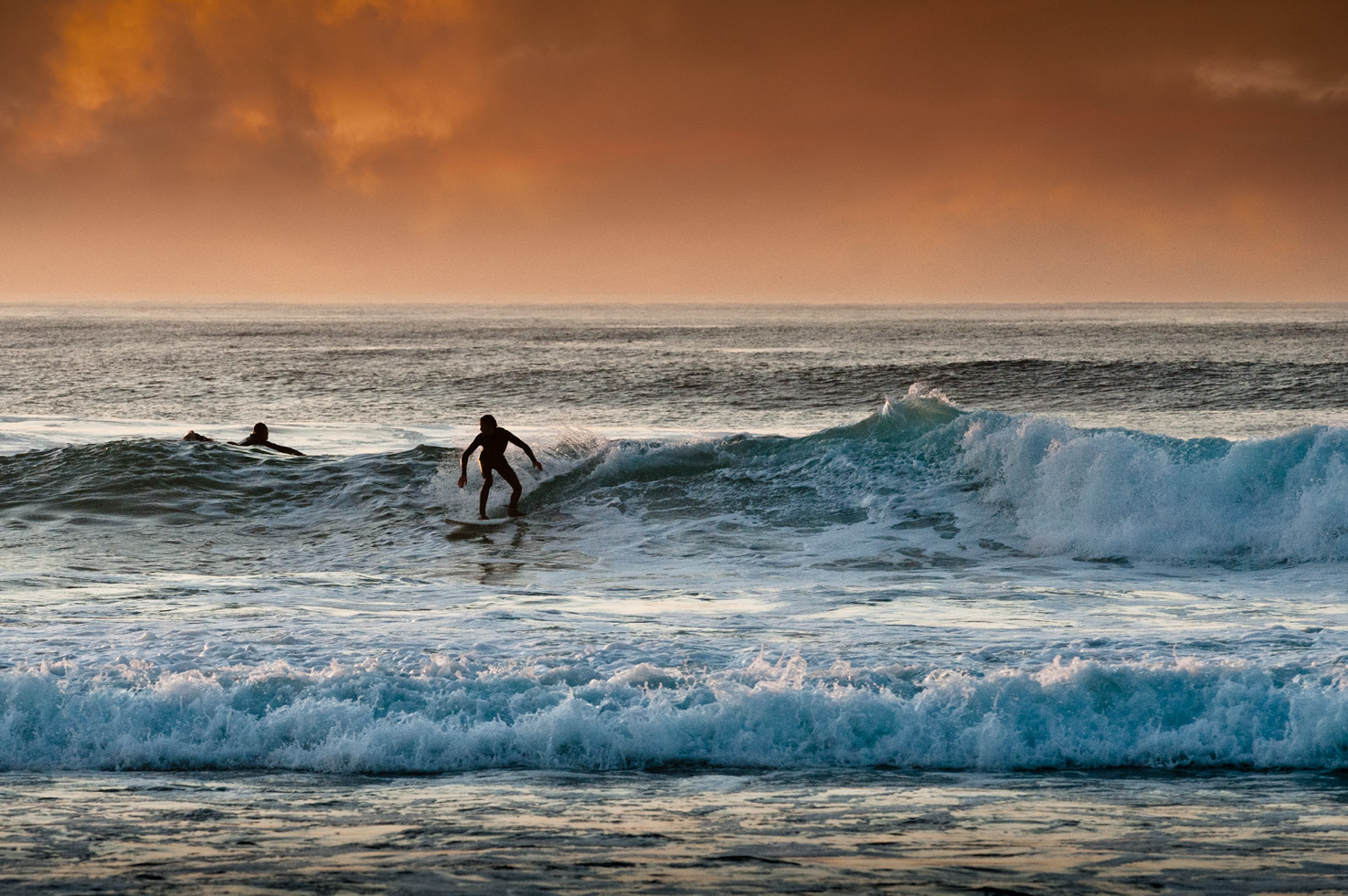 A surfer rides along a wave at sunset at Asilomar State Beach, Pacific Grove, California
