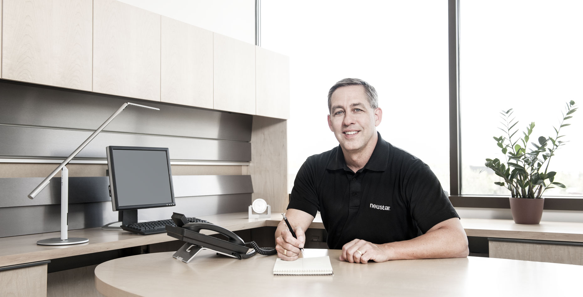 A man in a black polo shirt sitting at an office desk with a pen and pad of paper