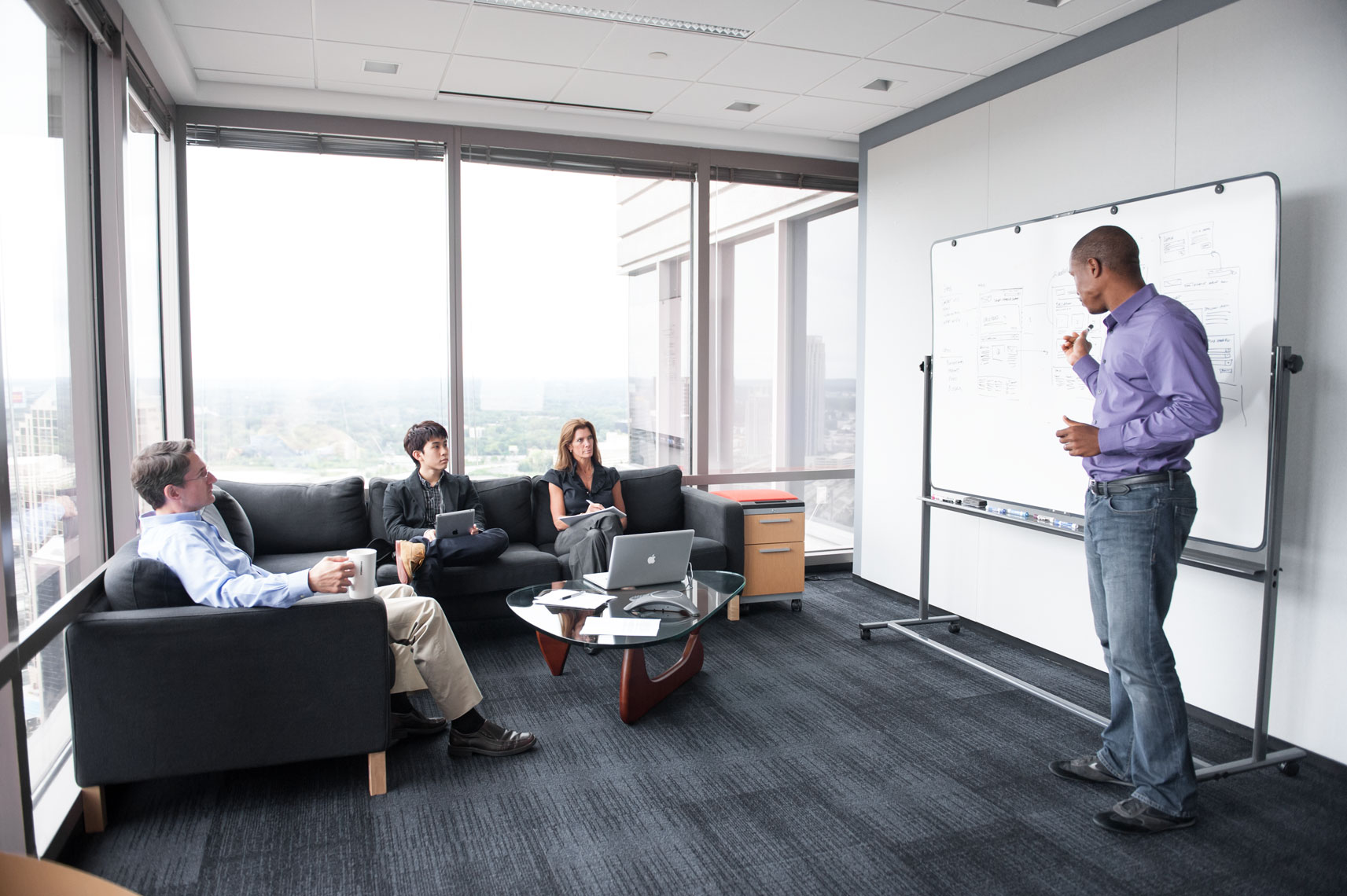 A man in a purple shirt standing at a whiteboard speaking to seated people in a high-rise office