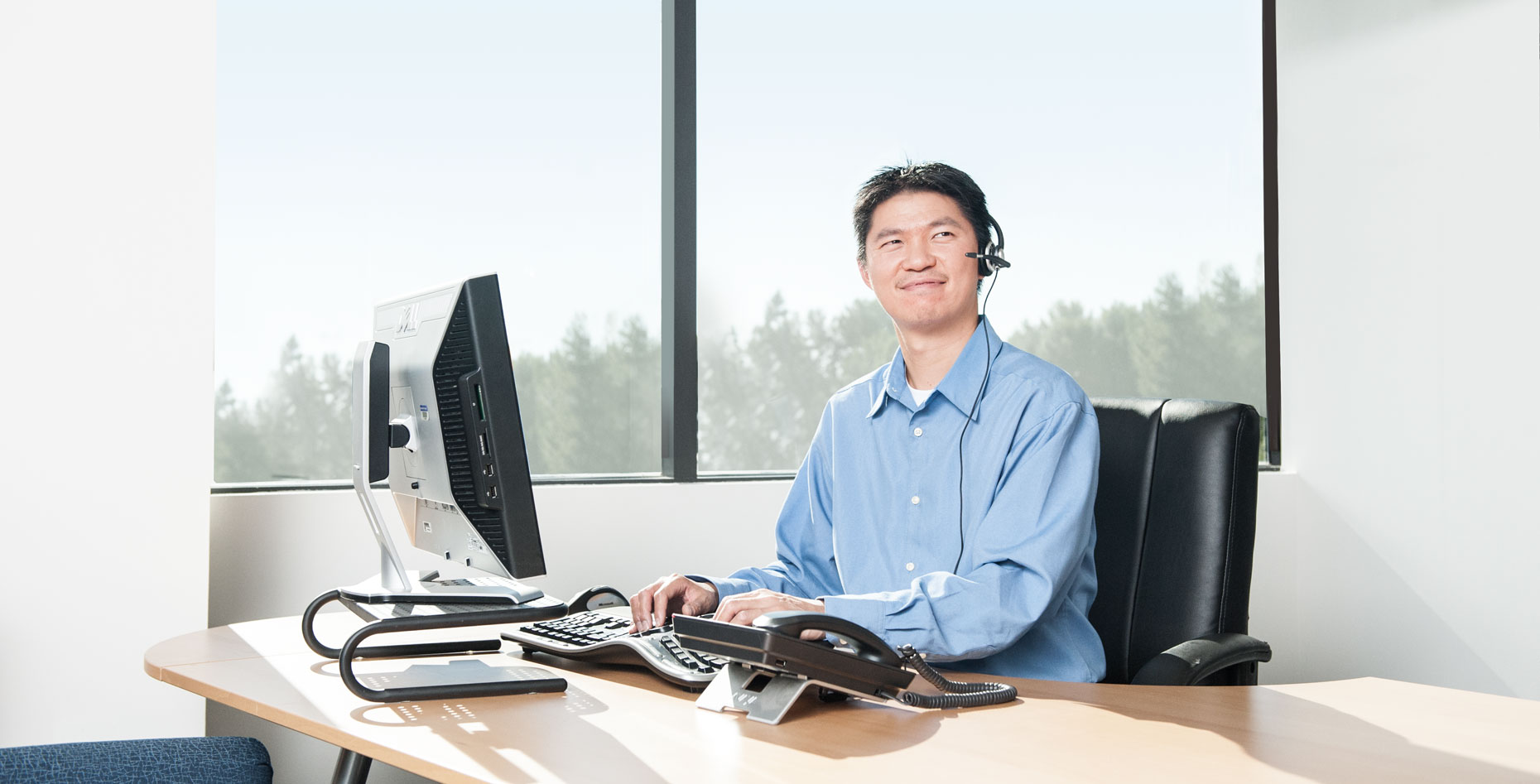A man in a blue shirt seated at a computer and wearing a hands-free phone headset