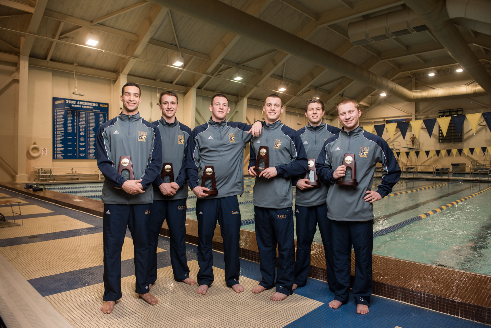 The TCNJ swim team holding their national championship plaques in front of a pool