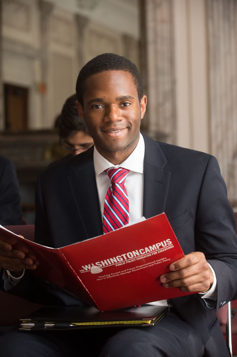 A student wearing a suit hold a red folder and smiles at the camera