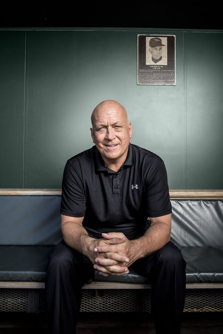 Cal Ripken seated in the Orioles dugout in Camden yards beneath a plaque honoring his father
