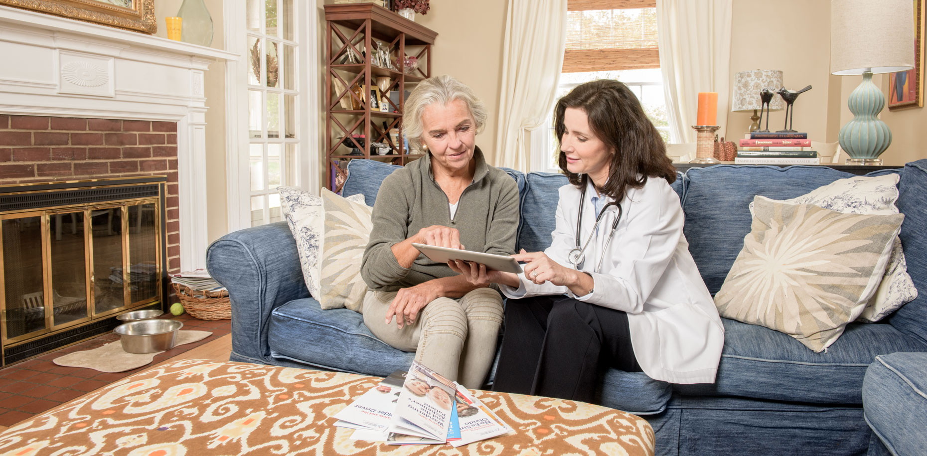 A nurse looks at computer tablet with a retiree on a sofa
