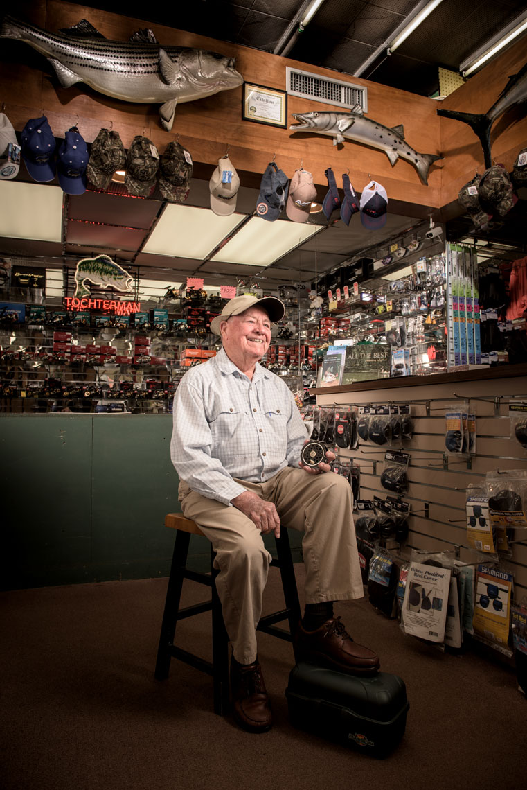 Lefty Kreh seated and holding a fly reel in front of the counter at Trochterman