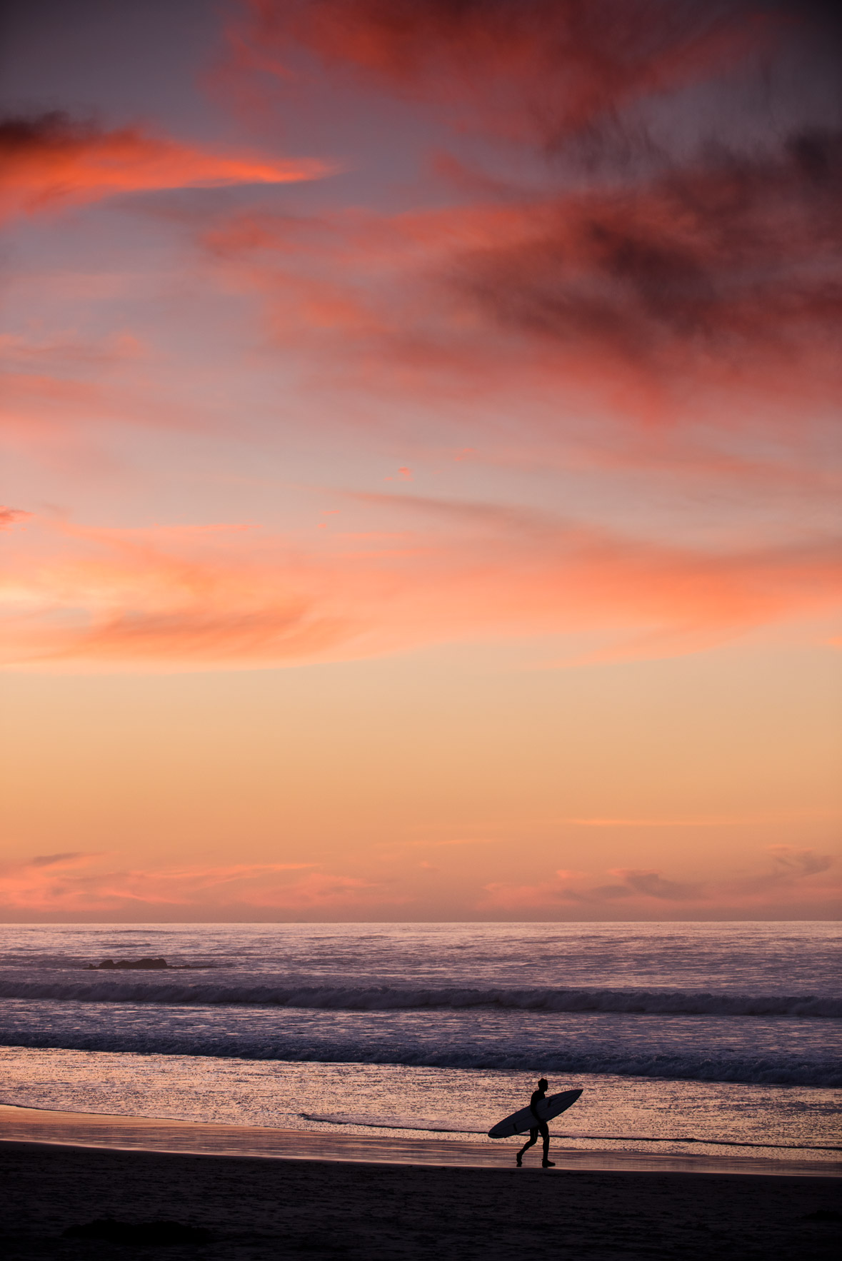 A surfer walks in silhouette under a sunset sky at Asilomar State Beach, Pacific Grove, California