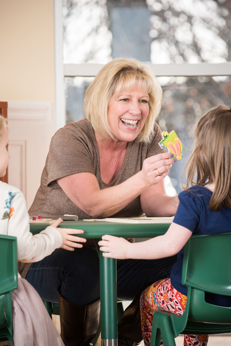 A smiling teacher shows seated kids a puzzle piece