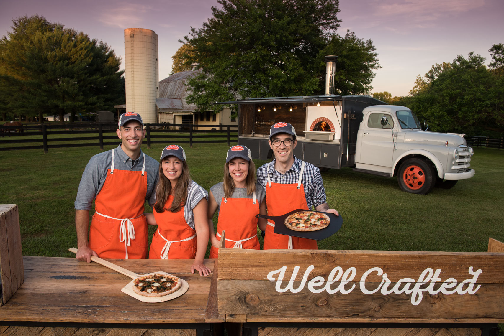 Four smiling pizza makers standing in front of their pizza truck on a farm