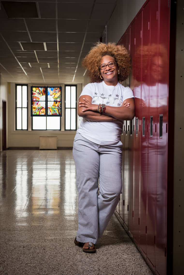 A high school principal leans against lockers in a darkened hallway with a student artwork in the background