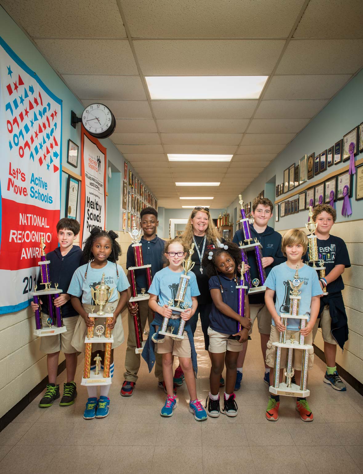 A group of chess champion elementary school students hold trophies with a teacher in a school hallway