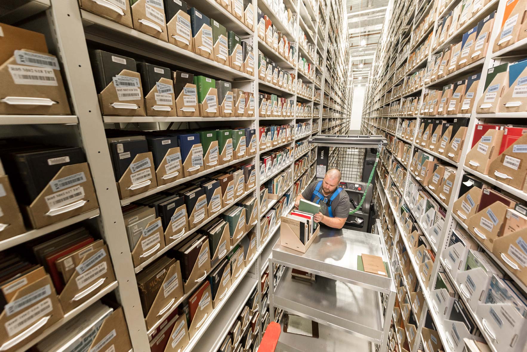 A man on a scissor-lift places books on a shelf in a large archive warehouse
