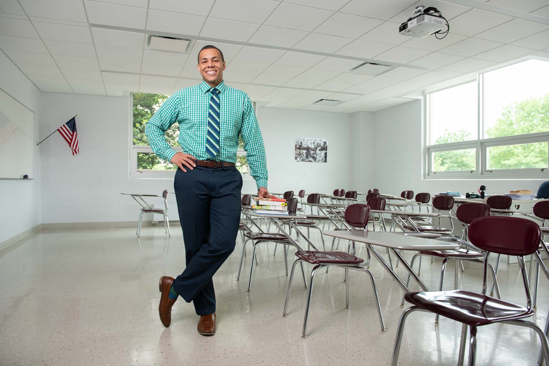 A teacher poses for the camera leaning on a desk in an empty classroom