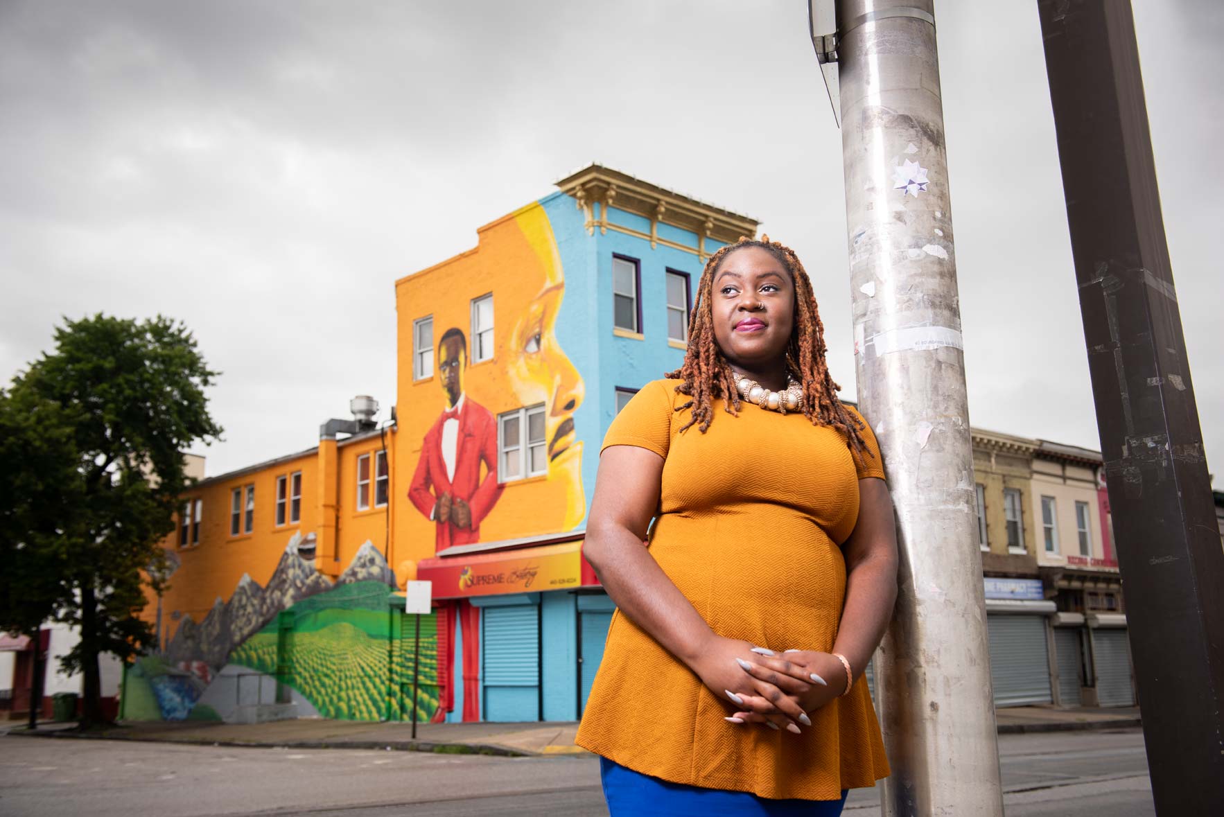 Lady Brion standing leaning against a post on a Baltimore street corner across from a colorful mural