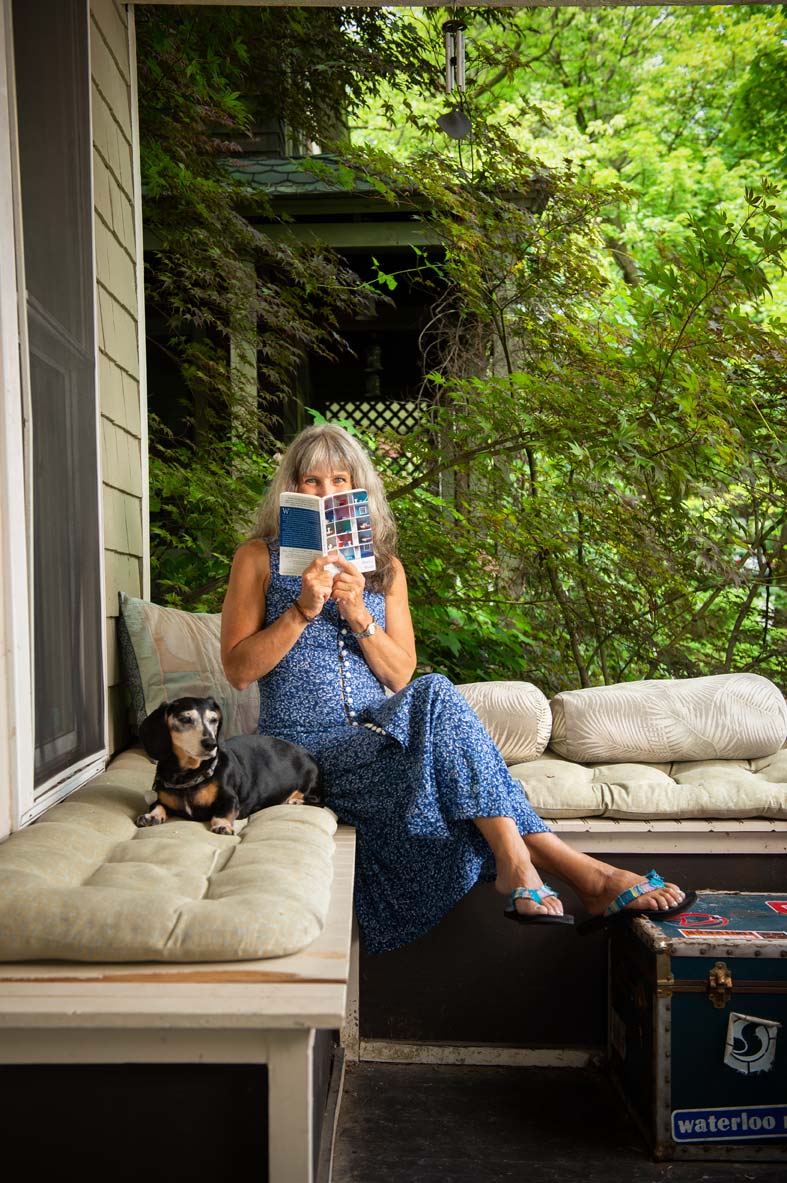Author Marion Winik seated on her porch and holding a book in front of her face