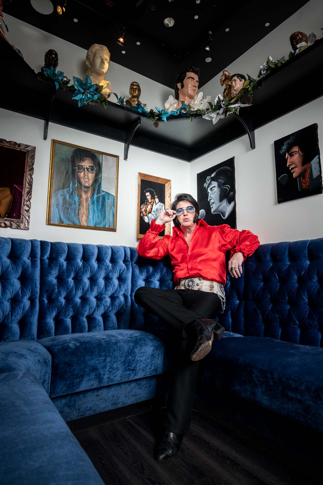 Elvis impersonator Jed Duvall wearing a red top and seated on a blue couch