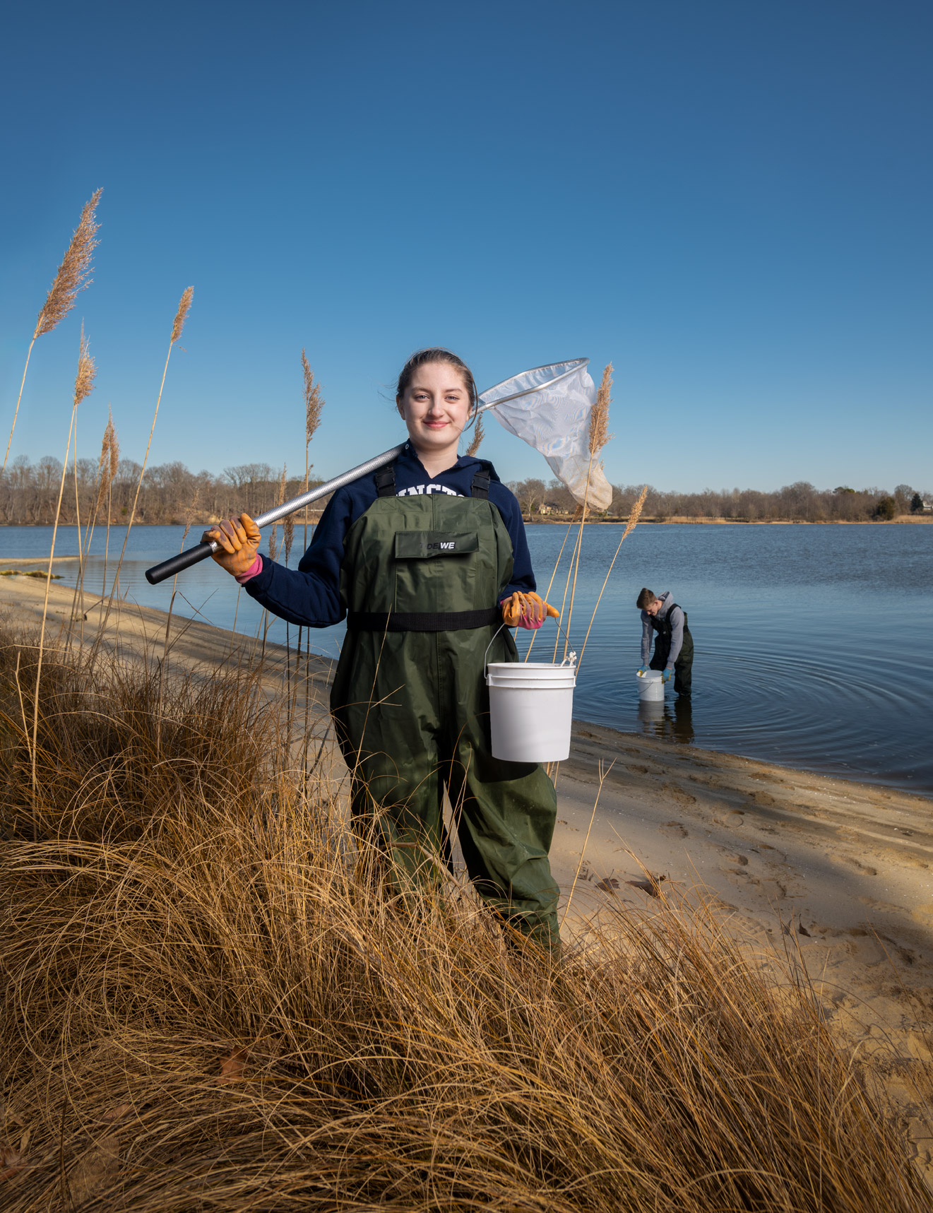 A high school student stands on the shoreline wearing waders and holding a net and a bucket