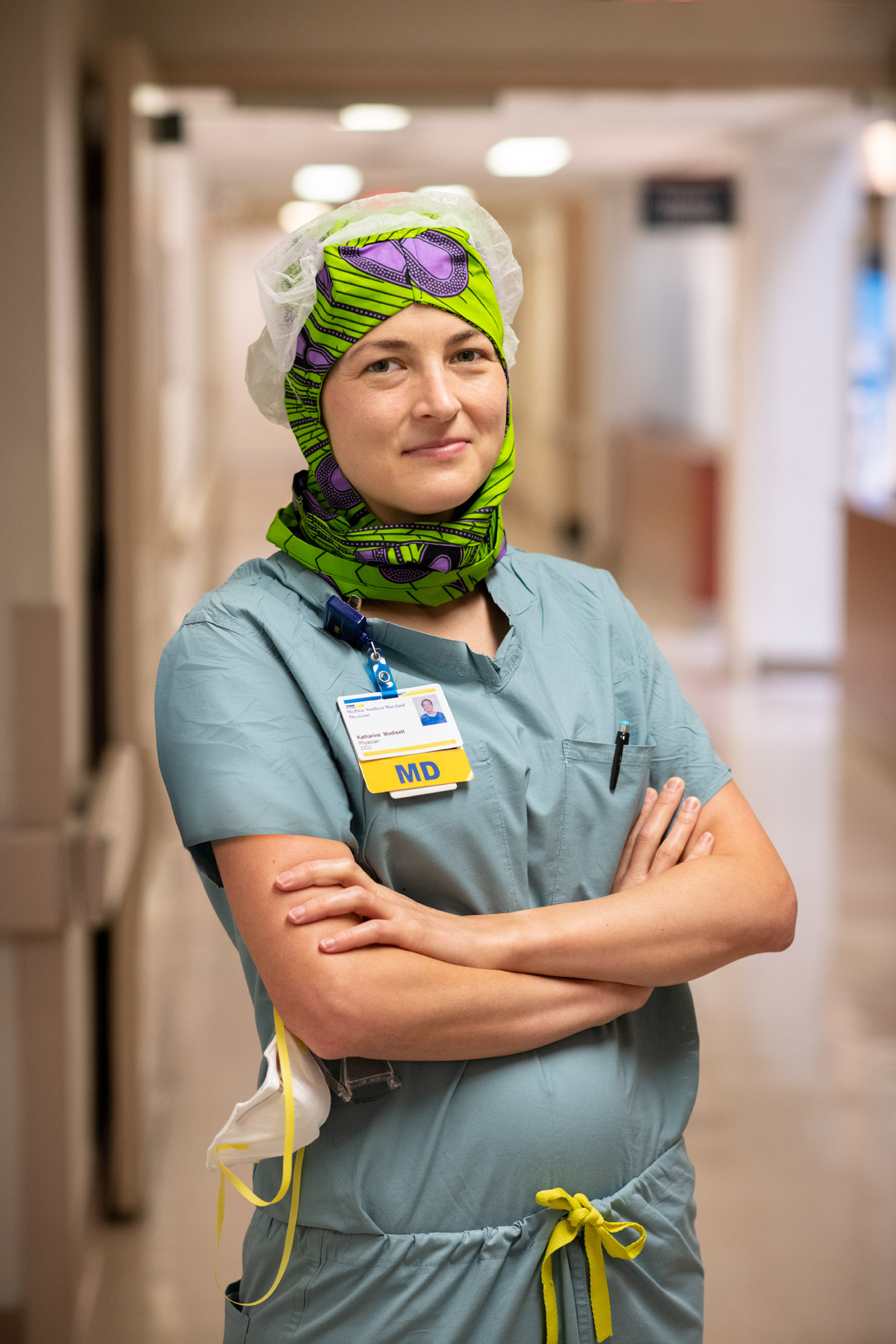 A hospital ICU doctor wearing scrubs standing with arms folded in a hallway